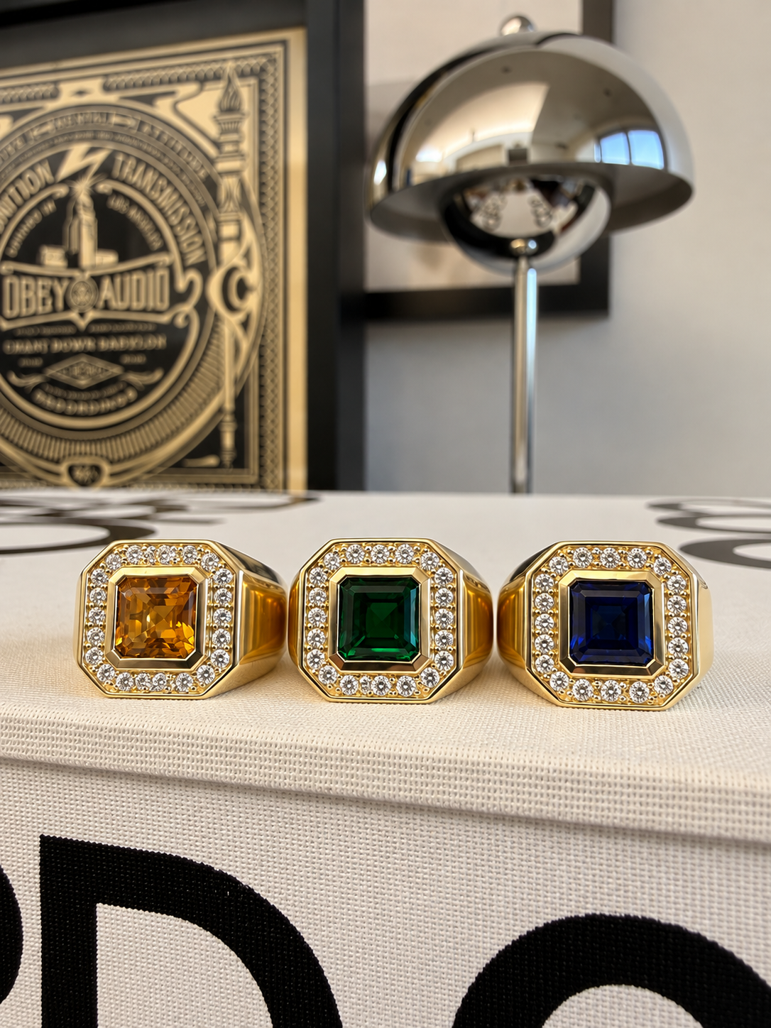 Three gold-toned ring stands with square gemstone tops are displayed on a white fabric surface. The left ring has a yellow-orange stone, the center has a green stone, and the right has a dark blue stone.